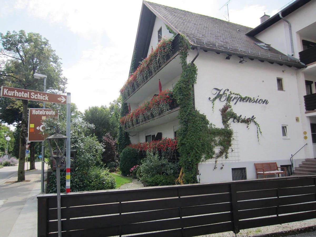 Hausecke mit efeubewachsener Fassade, blühenden Balkonkästen und Wegweiser zum Kurhotel Schick an der Straße