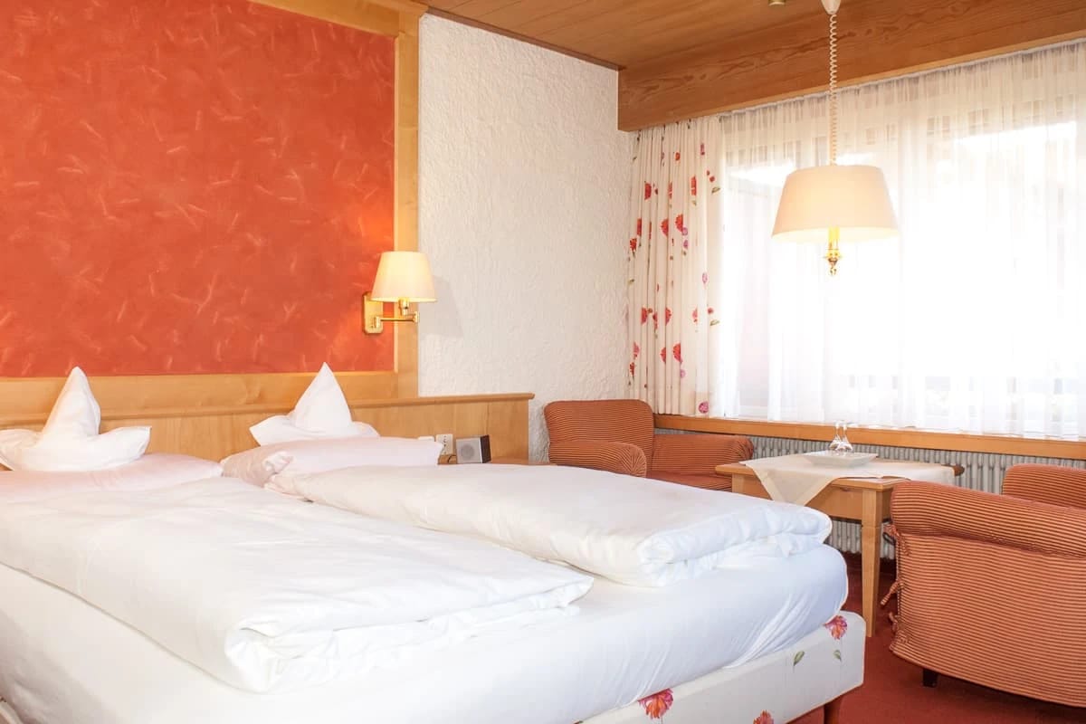 Doppelzimmer mit zwei Betten mit Holzkopfteil und Sesseln am Fenster