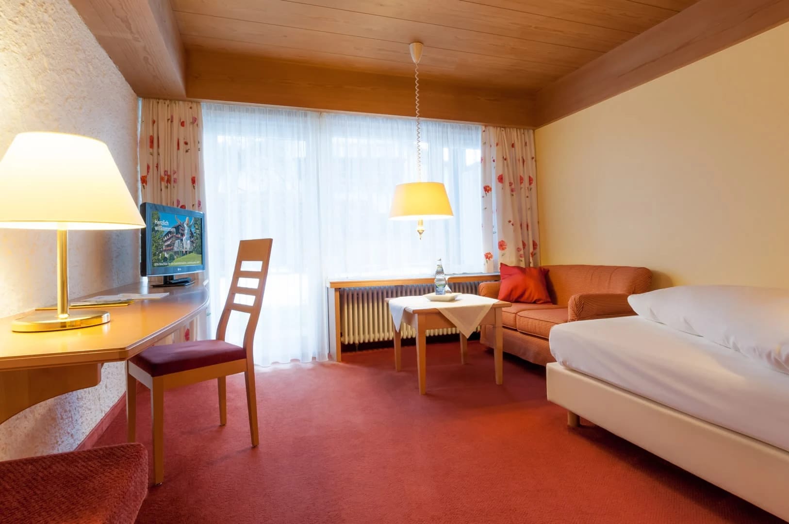Einzelzimmer mit Schreibtisch, Sofaecke, Bett und Holzdecke