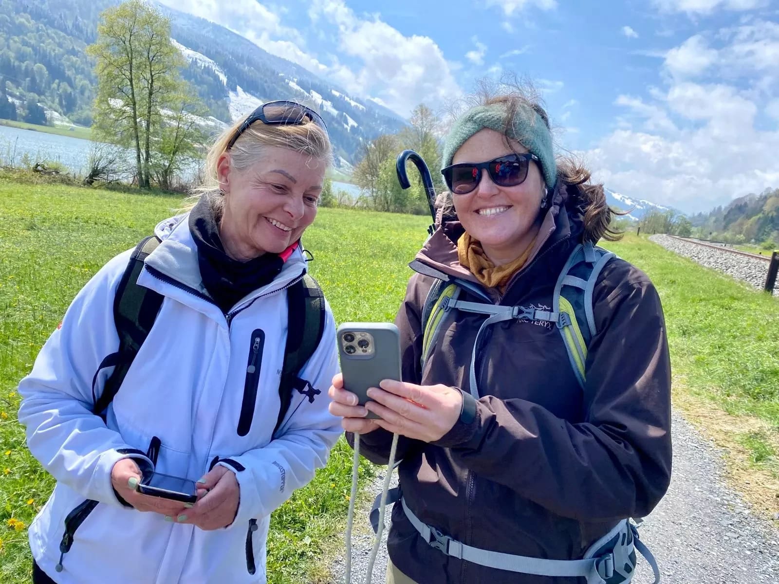 Zwei Wanderinnen schauen auf ein Smartphone vor Bergkulisse und See