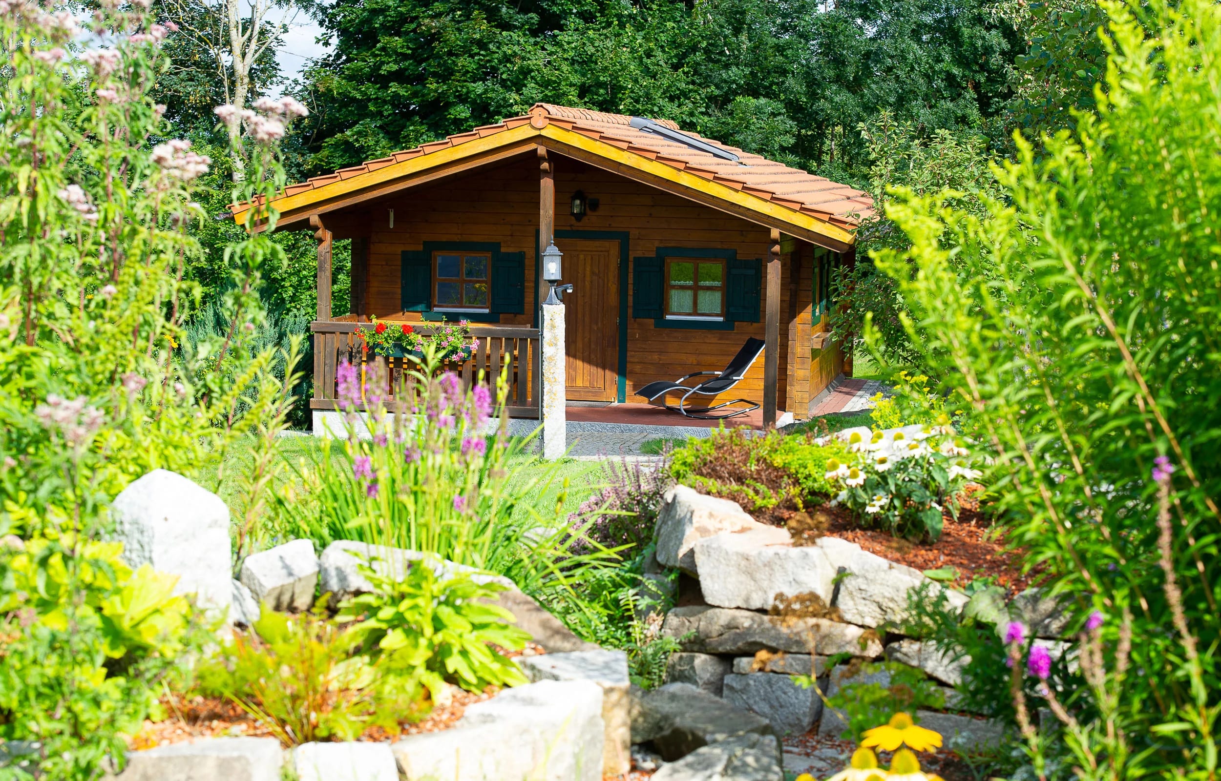 Kleines Holzhaus mit überdachter Veranda und Liegestuhl, umgeben von Staudenbeeten und Natursteinen