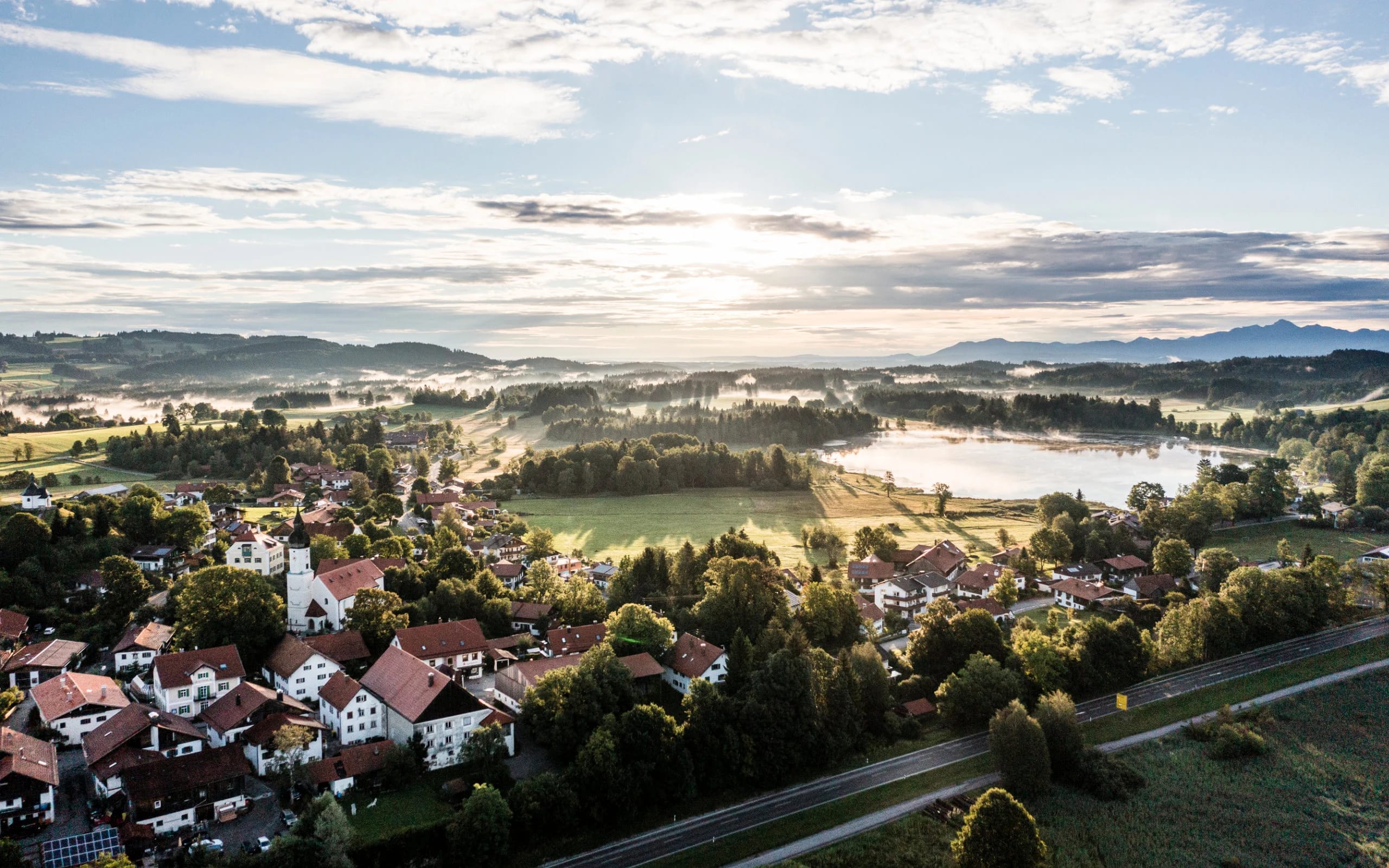 Luftaufnahme des Dorfes mit Kirche, Wiesen und dem See im Morgenlicht