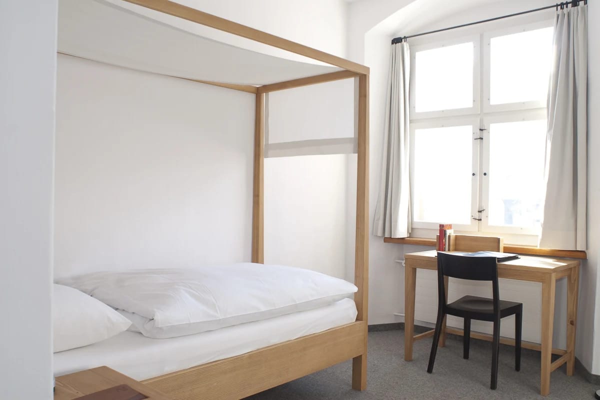 Schlichtes helles Zimmer mit hölzernem Bettgestell, weisser Bettwäsche und Schreibtisch am Fenster
