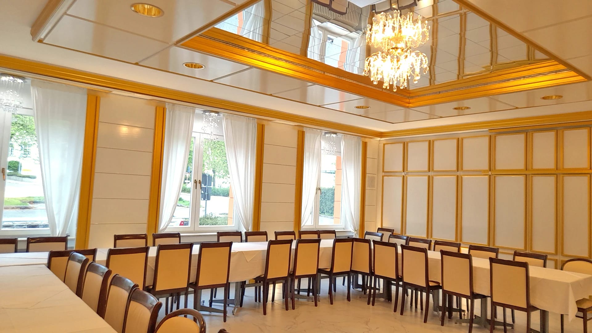 Speisesaal mit langen weiß gedeckten Tischen, Holzstühlen, Kronleuchter und hohen Fenstern