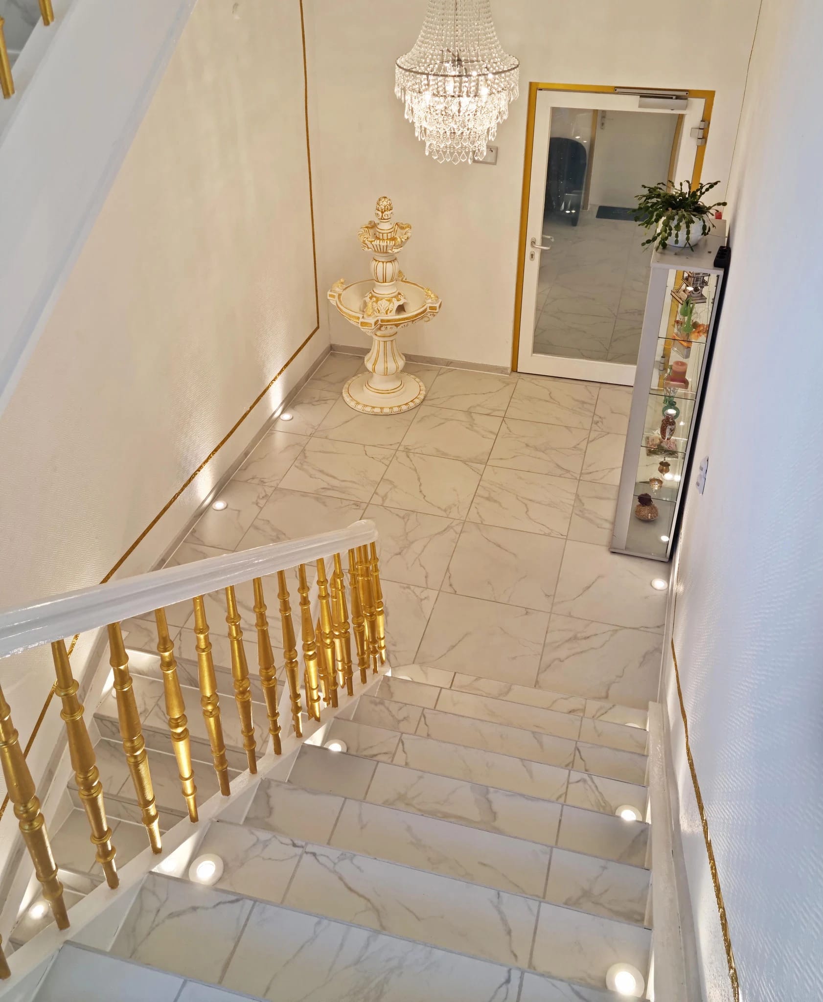Treppenhaus mit goldenem Geländer, Kronleuchter und hellem Marmorboden