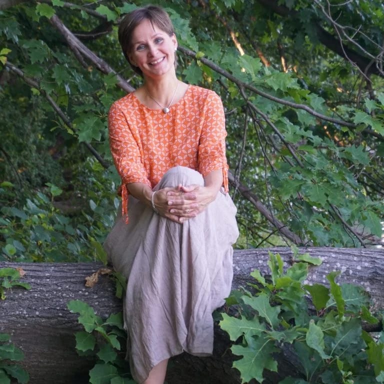 Diana sitzt auf einem Baumstamm im Wald, in einem orangefarbenen Oberteil