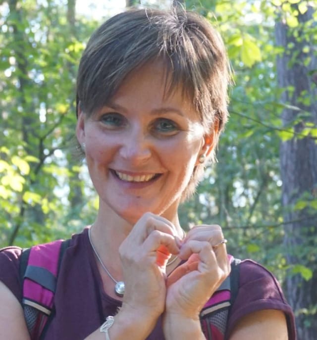 Porträt von Diana Baumgartl-Klemm, lächelnd mit einer Herzgeste vor herbstlichem Waldhintergrund