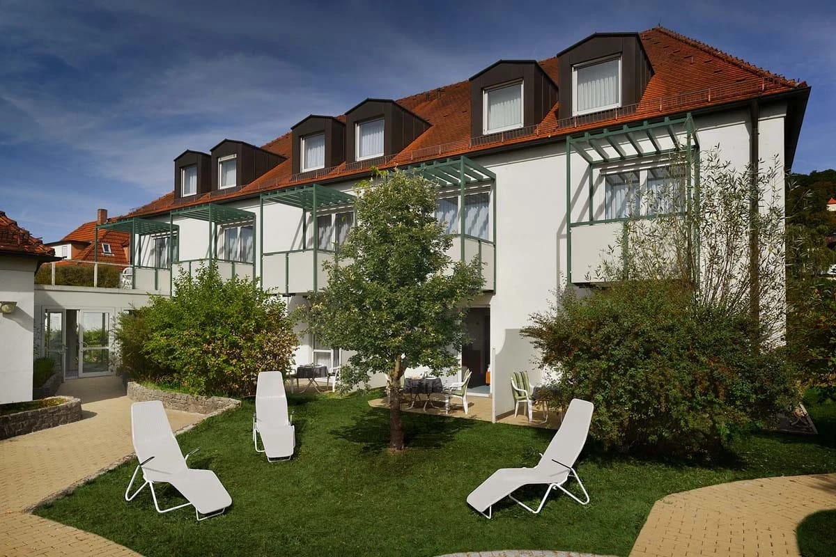 Weißes Hotelgebäude mit rotem Ziegeldach, Gauben und verglasten Balkonen, davor Liegestühle auf der Rasenfläche