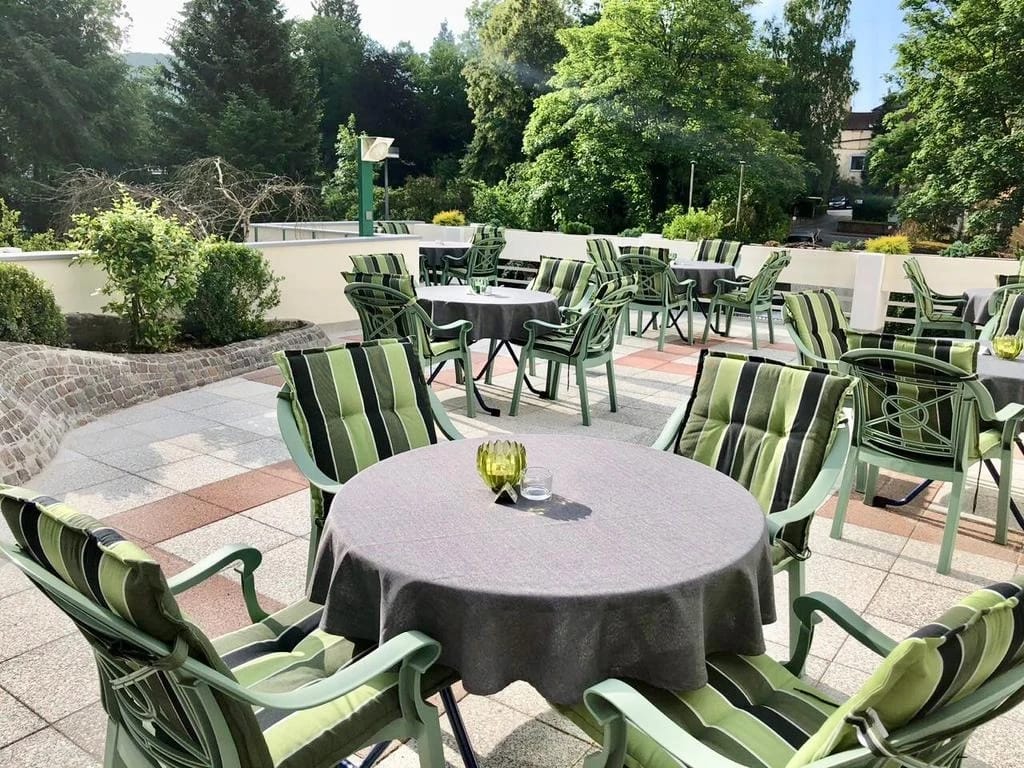 Sonnige Terrasse mit runden Tischen und grün gestreiften Stühlen, umgeben von Bäumen