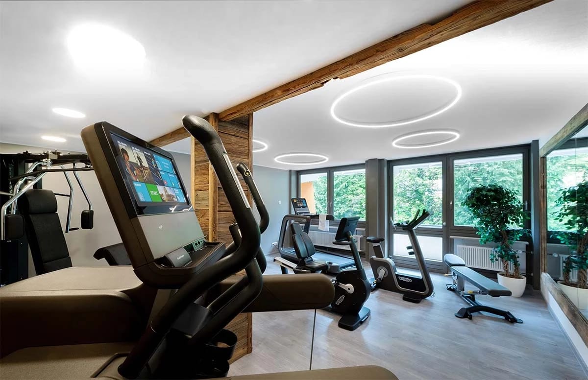 Fitnessraum mit Ergometern und Kraftgeräten vor bodentiefen Fenstern ins Grüne