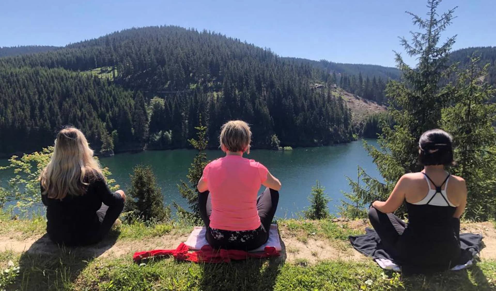 Drei Frauen sitzen zur Meditation auf einer Wiese oberhalb eines von Wald umgebenen Sees