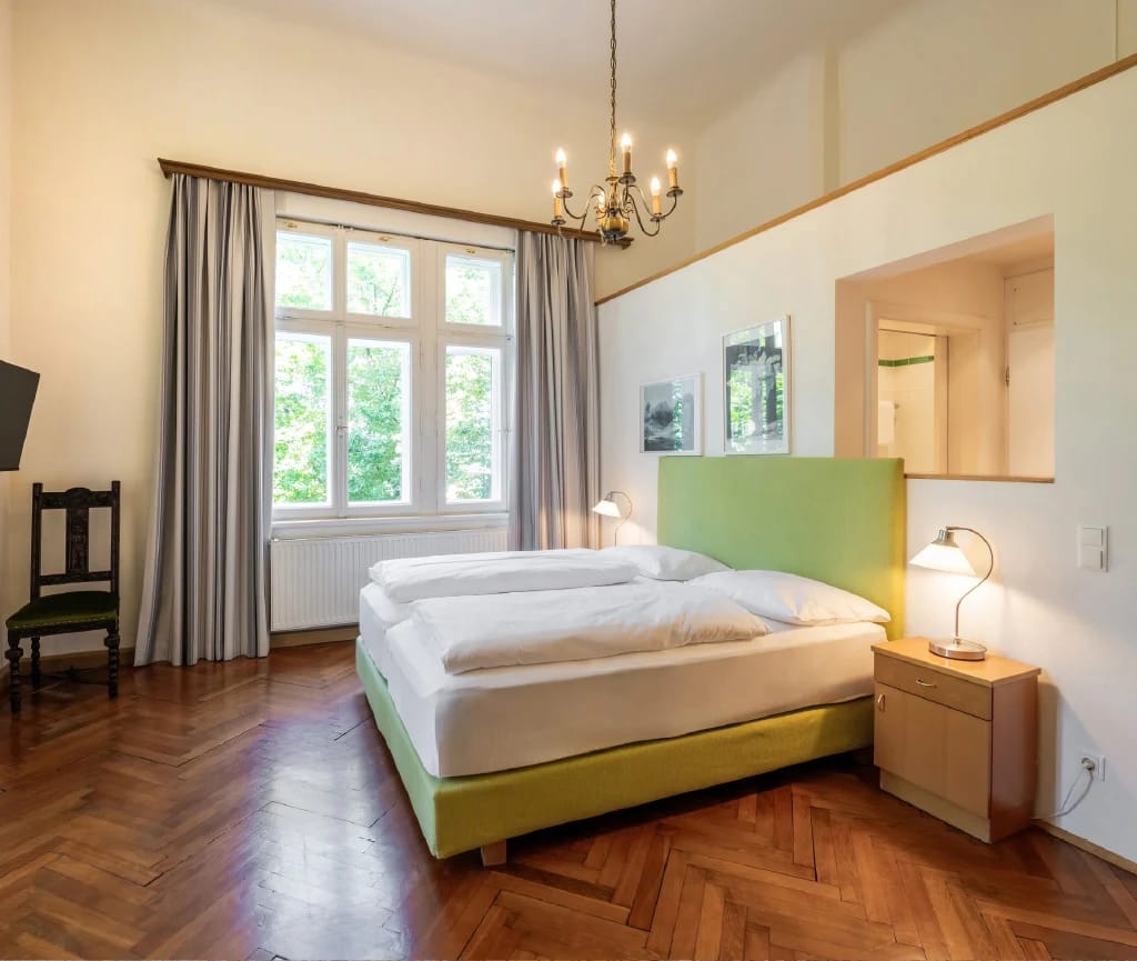 Doppelzimmer mit Fischgrätparkett, Sprossenfenster, Luster und Bett mit grünem Kopfteil