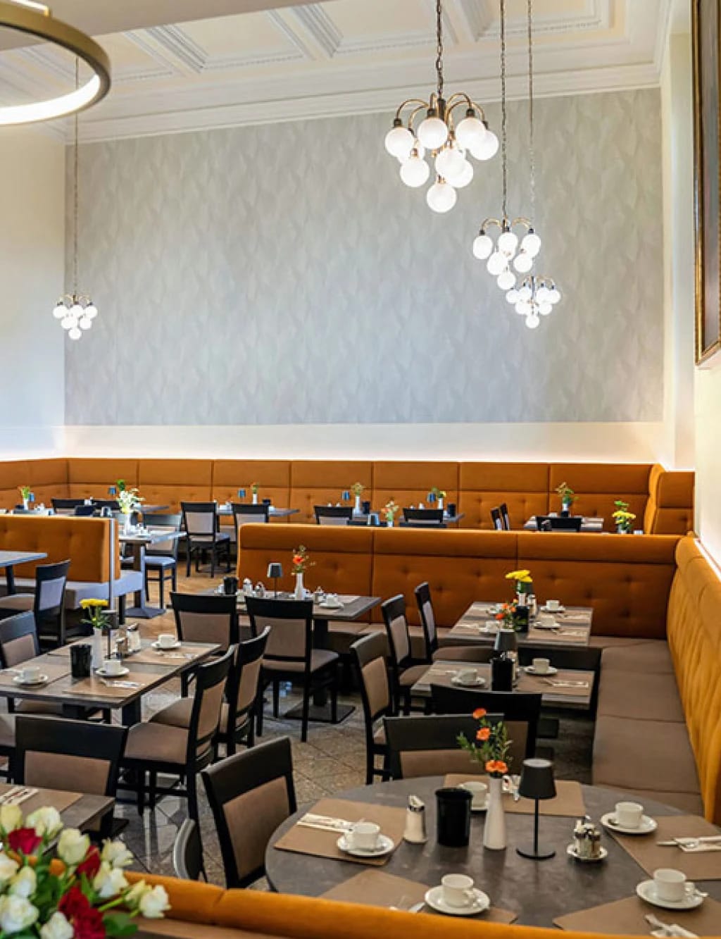 Speisesaal mit orangefarbener Sitzbank, eingedeckten Tischen und Kugelleuchten unter hoher Decke