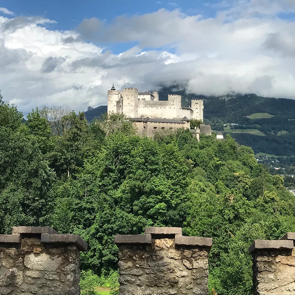 Blick von einer Steinmauer über bewaldeten Hang auf die Festung Hohensalzburg