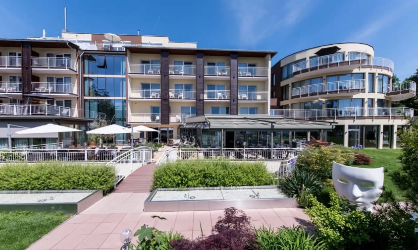 Außenansicht des Hotels mit Balkonen, verglastem Rundbau und Gartenanlage bei Sonnenschein