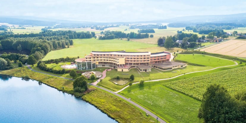 Luftaufnahme des Hotelgebäudes am Seeufer, umgeben von Wiesen und Wald