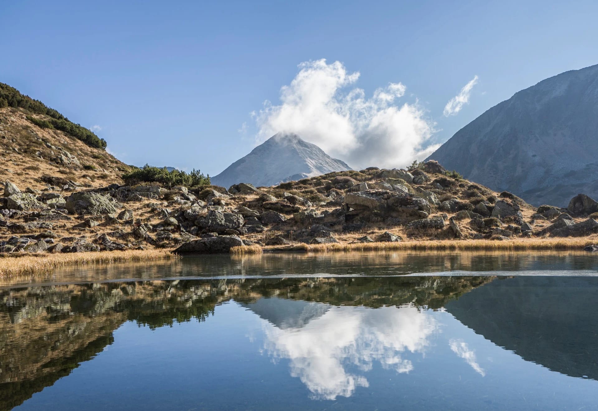 Bergsee mit Spiegelung einer Wolke, felsigem Ufer und Gipfel dahinter