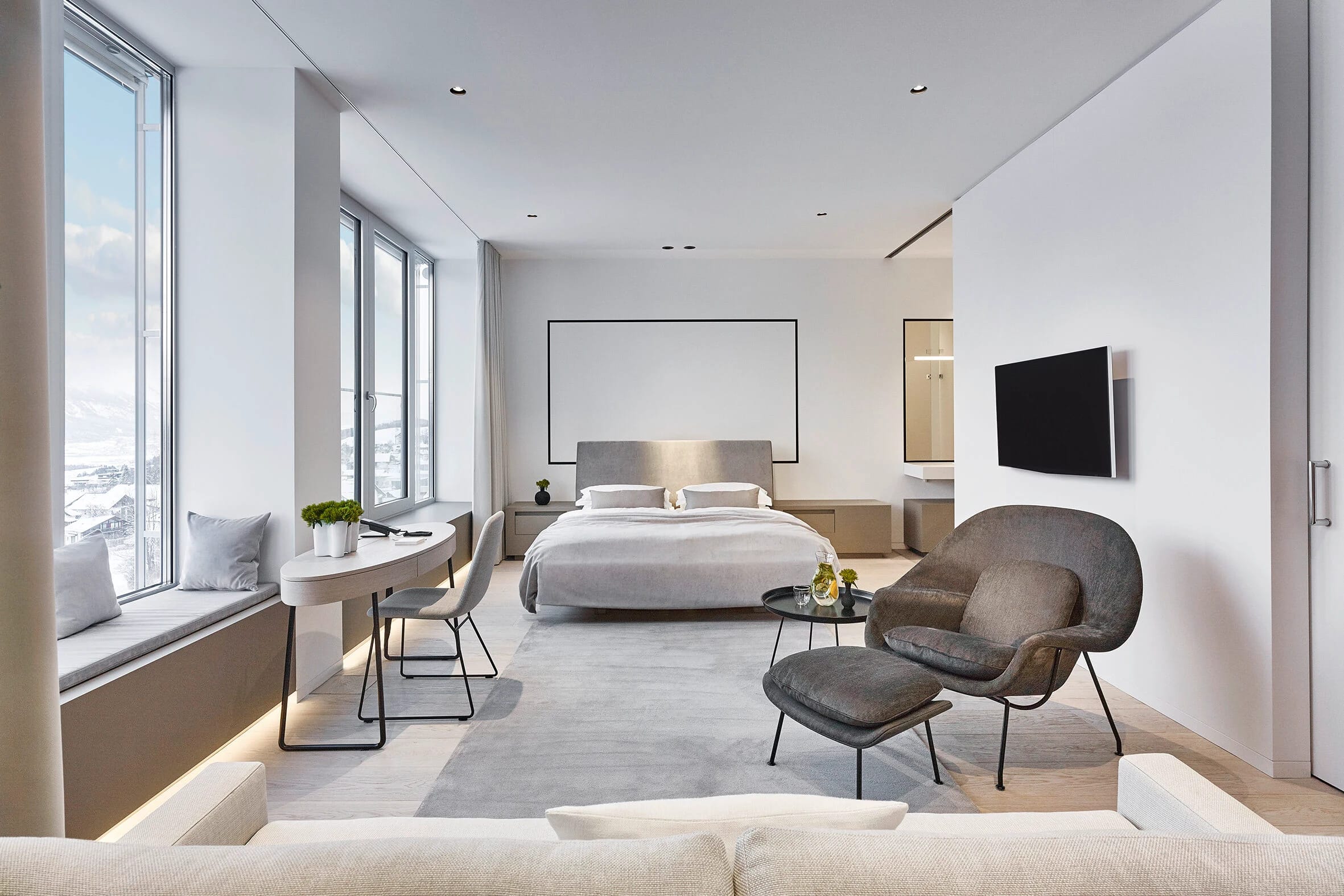 Helle Suite mit Bett, Schreibtisch an der Fensterfront und Sessel mit Hocker, Blick auf verschneite Berge