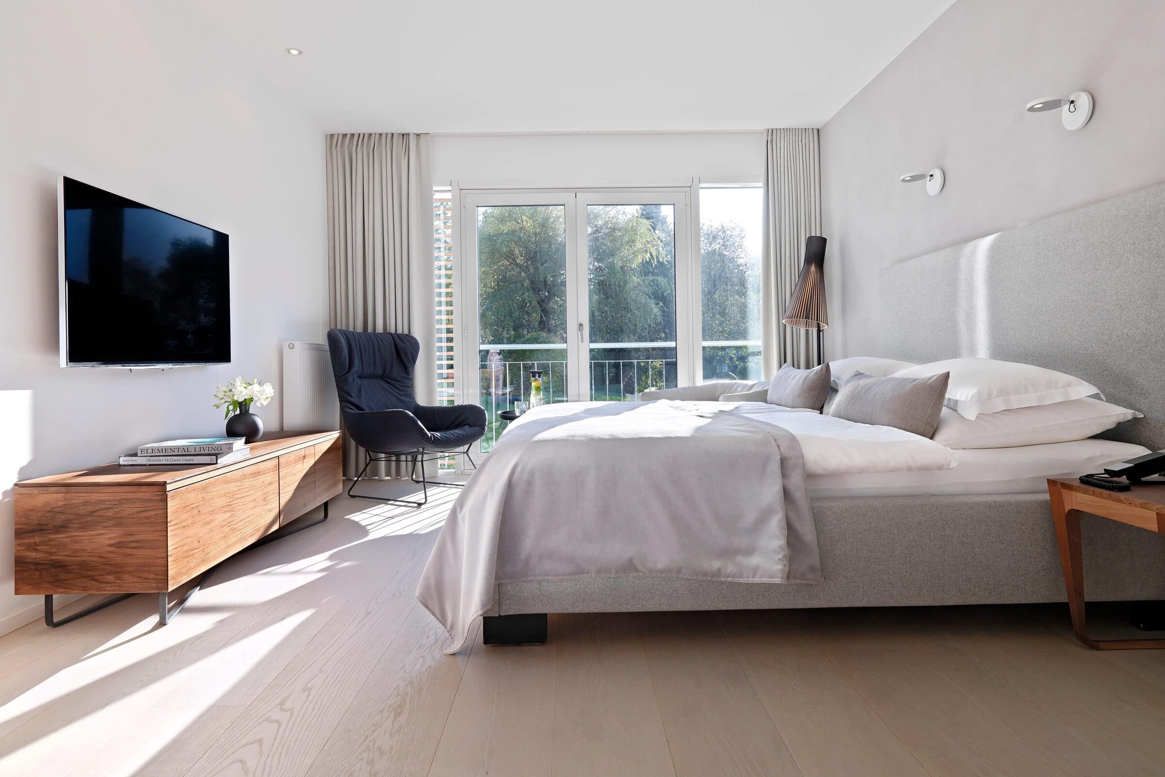 Doppelzimmer mit Bett, Sideboard aus Holz und dunkelblauem Sessel an der Balkontür