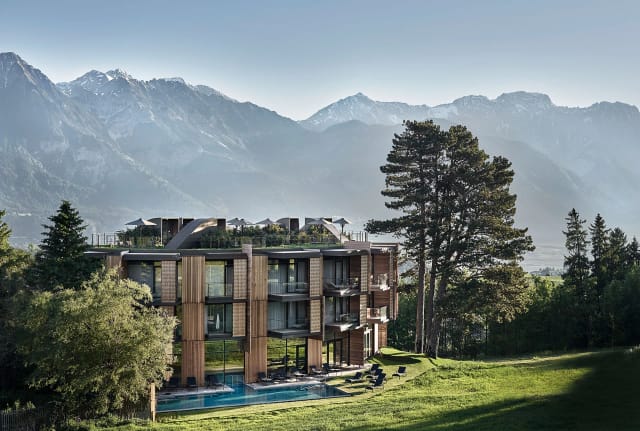 Außenansicht des Hauses mit Holzfassade und begrünten Dachterrassen, davor der Außenpool, dahinter die Berge
