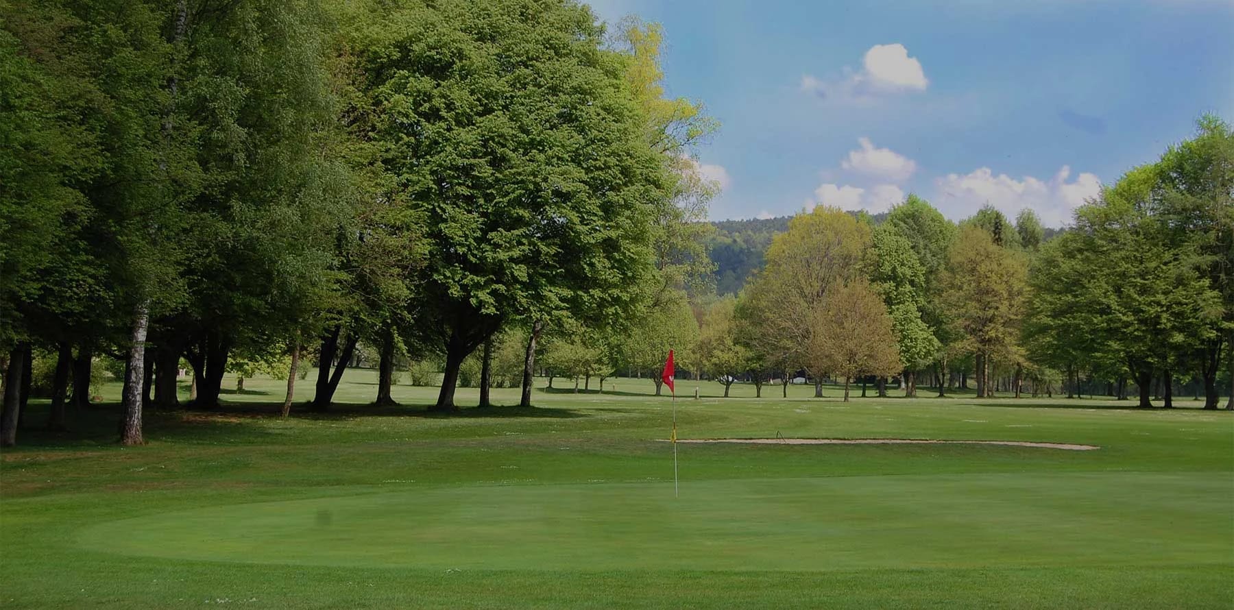 Golfplatz mit grünem Rasen, Bäumen und Hügeln unter blauem Himmel