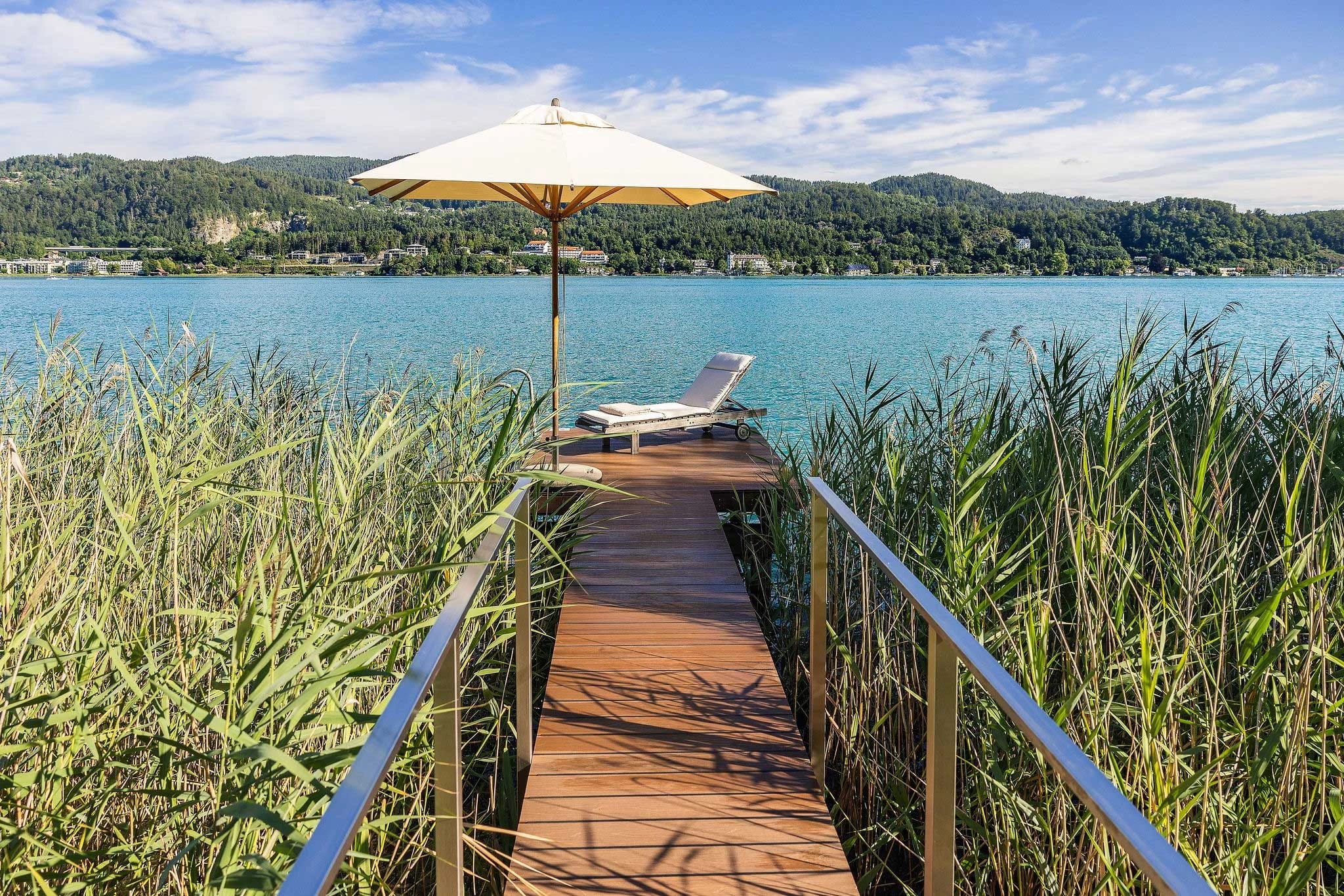 Holzsteg durch das Schilf mit Liege und Sonnenschirm am Seeufer