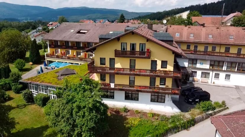 Luftaufnahme des gelben Hotelgebäudes mit Balkonen, Außenpool und Garten vor bewaldeten Hügeln