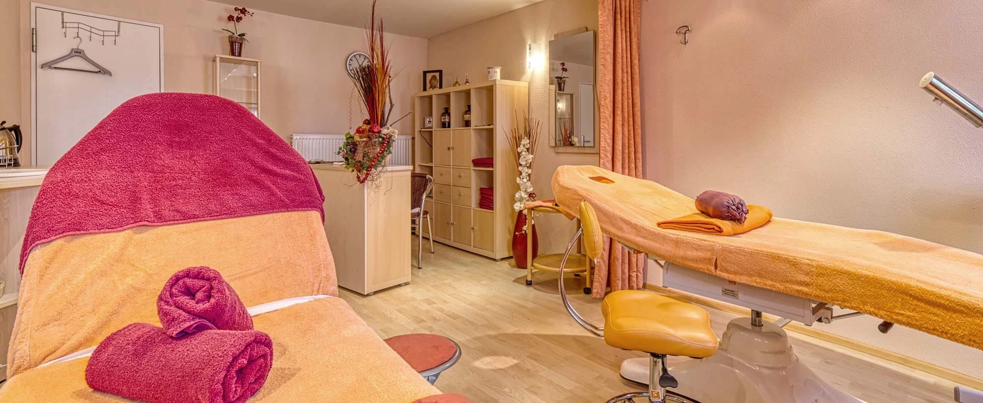 Behandlungsraum mit zwei Massageliegen, gerollten Handtüchern und Regal