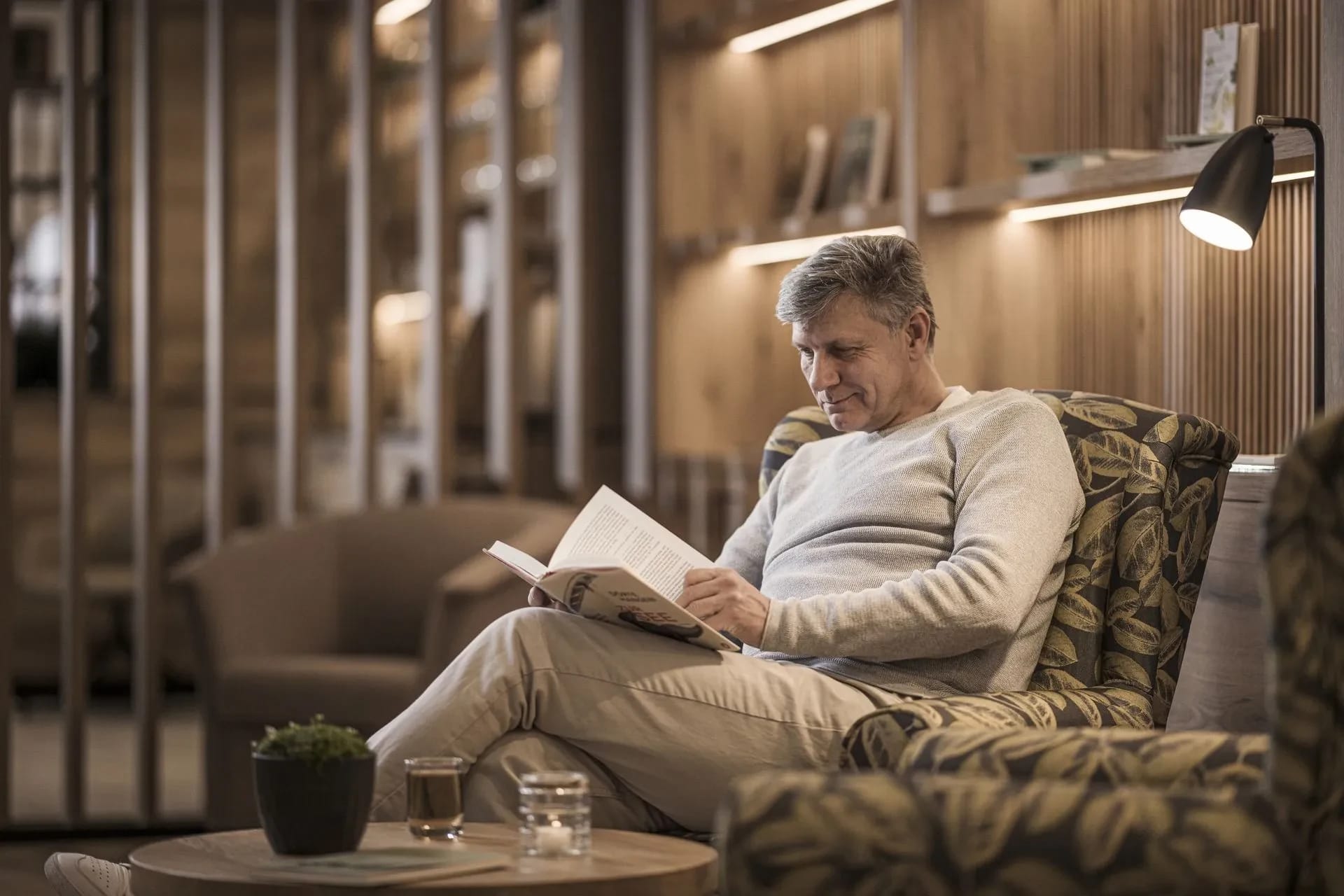 Lounge mit Sessel, Stehlampe und beleuchtetem Holzregal, ein Gast liest ein Buch