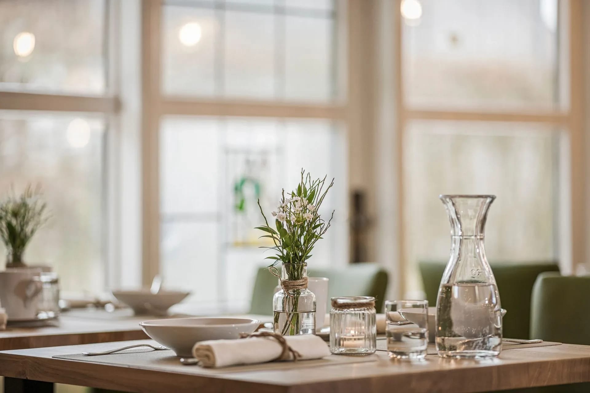 Gedeckter Tisch im Restaurant mit Wasserkaraffe, Gläsern und Blumenvase vor Sprossenfenstern