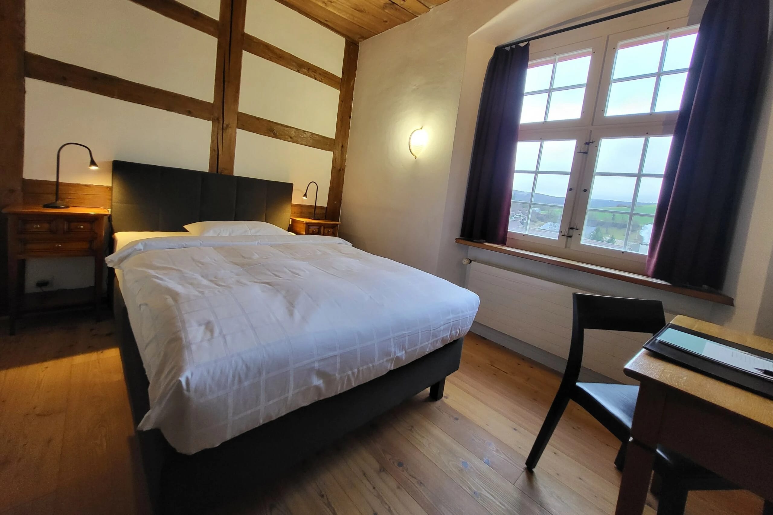 Doppelzimmer mit Fachwerkwand, dunklem Bettkopfteil und grossem Fenster mit Blick ins Grüne