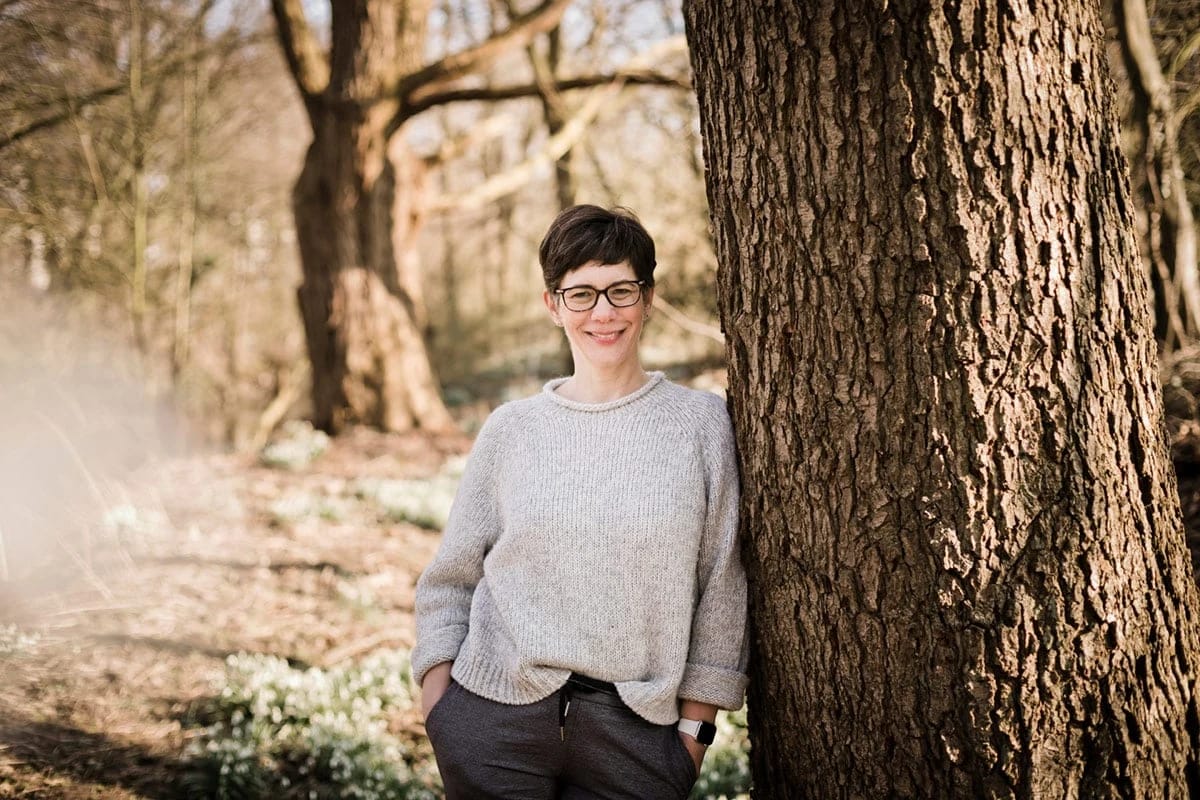 Dr. Nicola Stein steht lächelnd mit Brille an einem großen Baumstamm im Wald