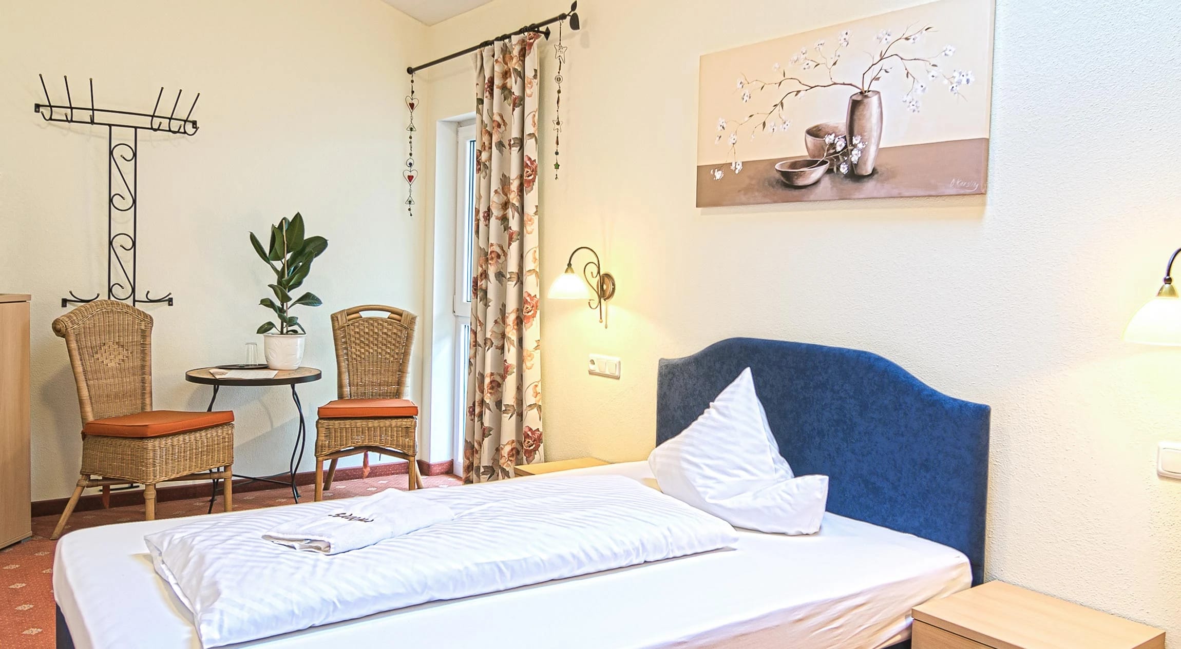 Einzelzimmer mit Bett, Rattan-Sitzecke am Fenster und heller Einrichtung