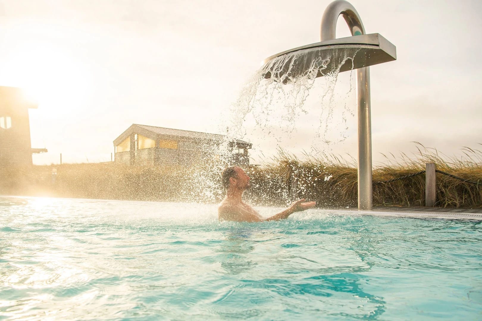 Außenbecken der benachbarten Dünen-Therme mit Schwalldusche zwischen Dünengras im Abendlicht