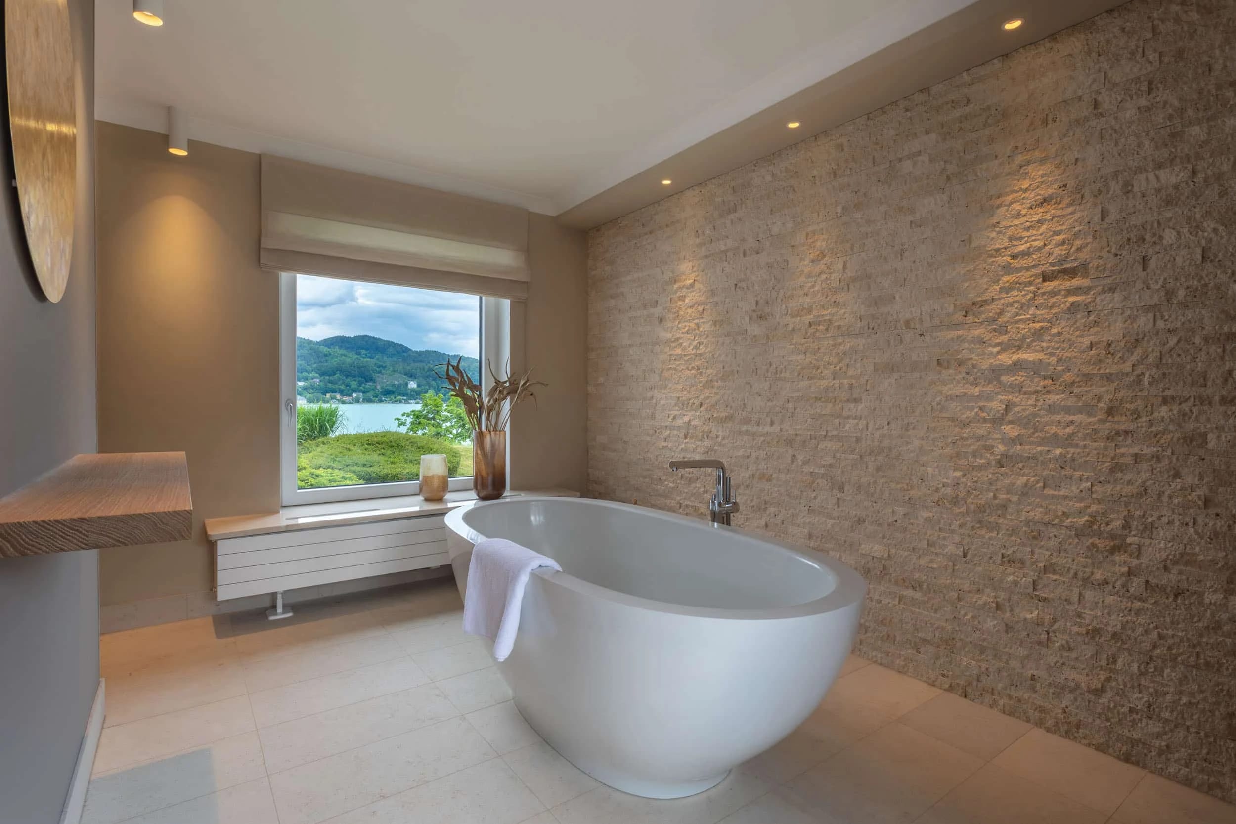 Badezimmer mit freistehender Badewanne, Natursteinwand und Fenster zum See