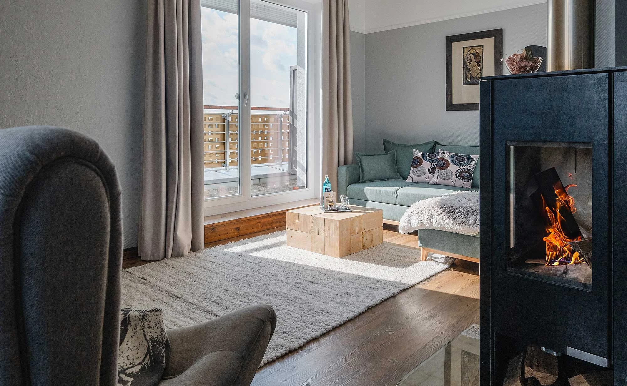 Wohnbereich einer Suite mit brennendem Kaminofen, Sofa, Sessel und bodentiefem Fenster