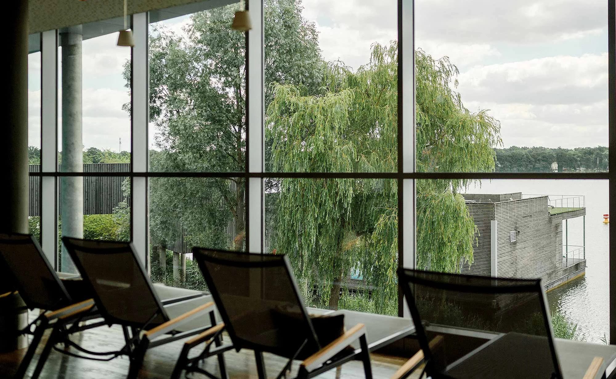 Ruhebereich mit dunklen Liegen vor einer raumhohen Glasfront mit Blick auf Weiden und den See