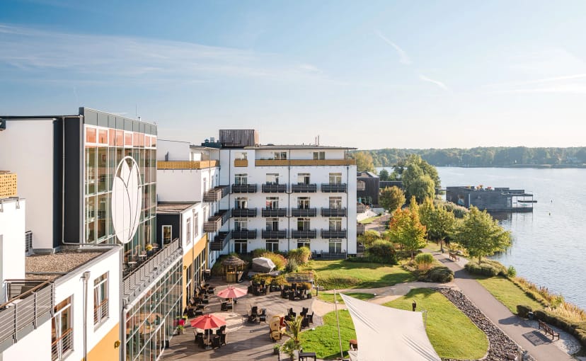 Hotelgebäude mit weißer Fassade und Balkonen, davor eine Terrasse mit roten Sonnenschirmen, rechts der Ruppiner See