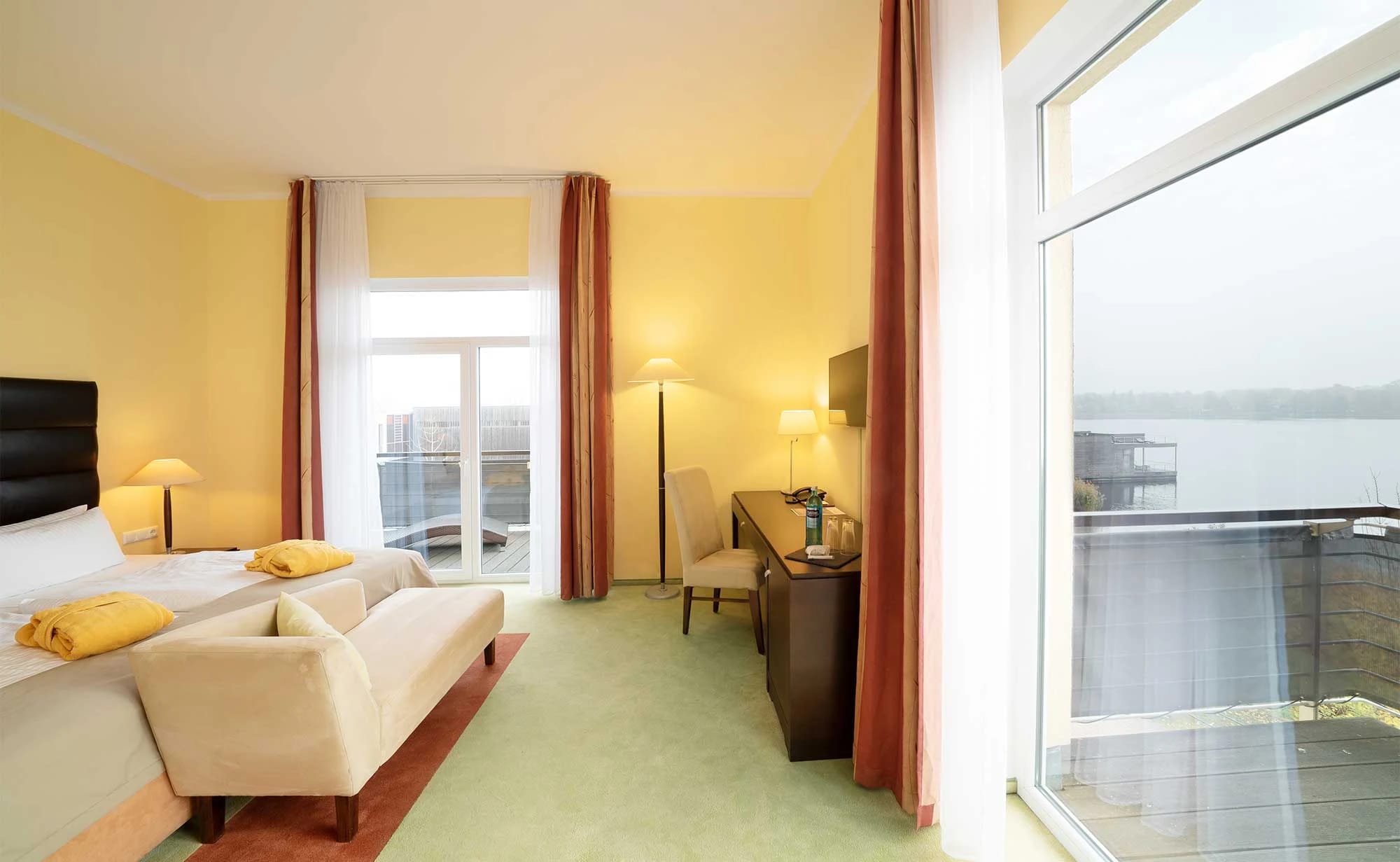 Eckzimmer mit Doppelbett, Sitzecke und Schreibtisch vor bodentiefen Fenstern mit Blick auf den See