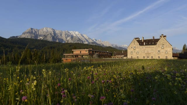 Steinernes Haupthaus mit angebautem Holztrakt vor schroffen Felsbergen, davor eine blühende Bergwiese