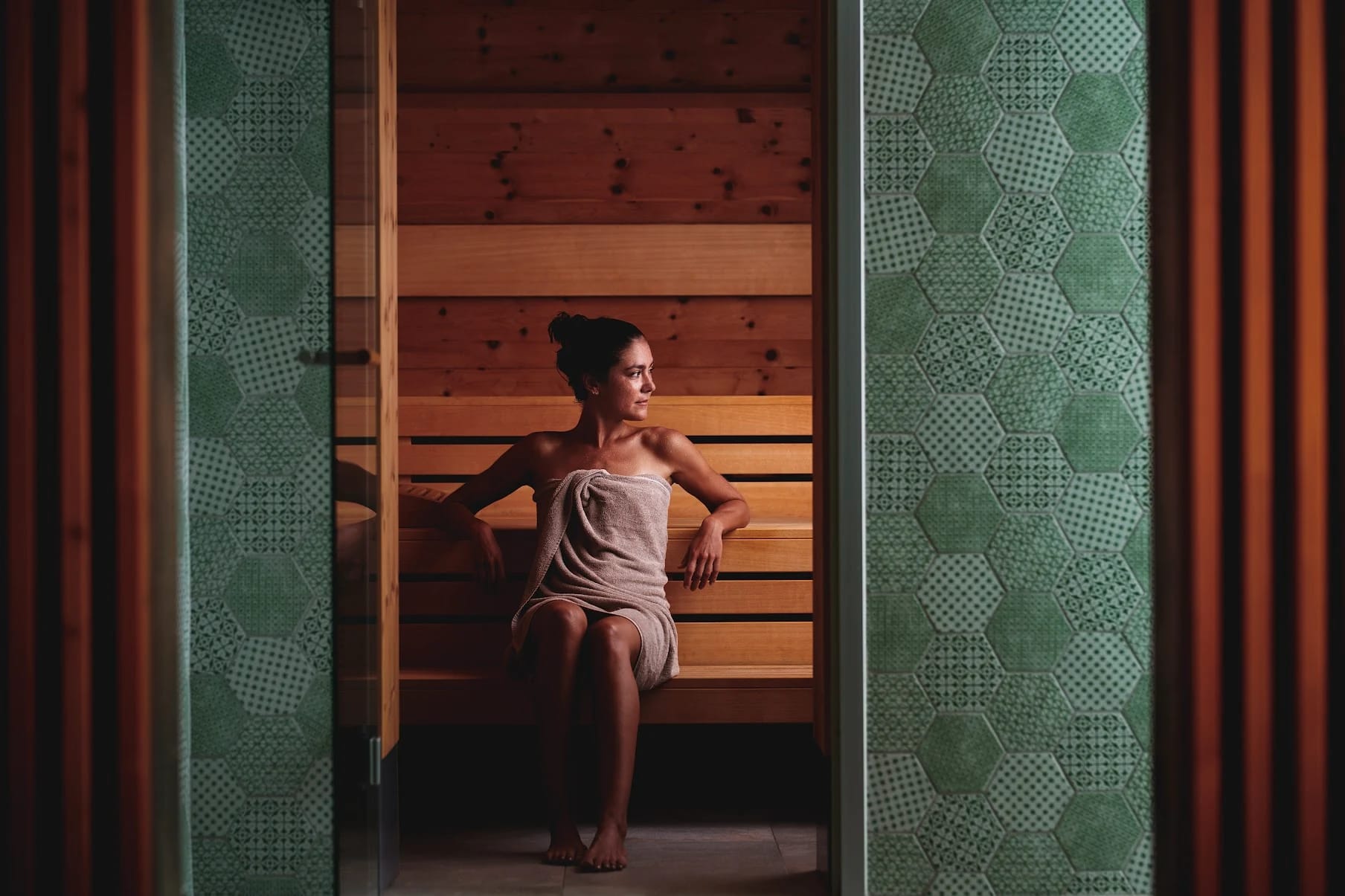 Sauna aus hellem Holz mit Holzbänken, Blick durch einen grün gefliesten Türrahmen