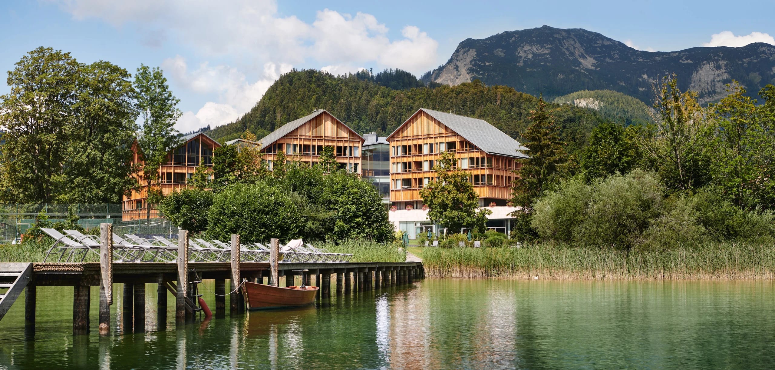 Blick über den See auf die Häuser des Resorts mit Steg, Liegestühlen und Holzboot