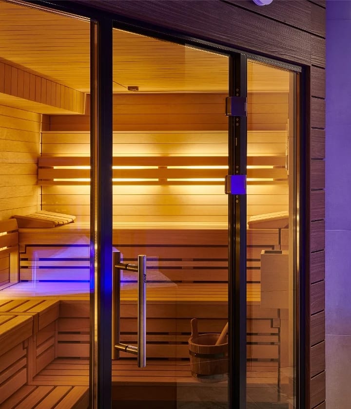 Warm beleuchtete Sauna mit Holzbänken und Holzkübel hinter einer Glastür
