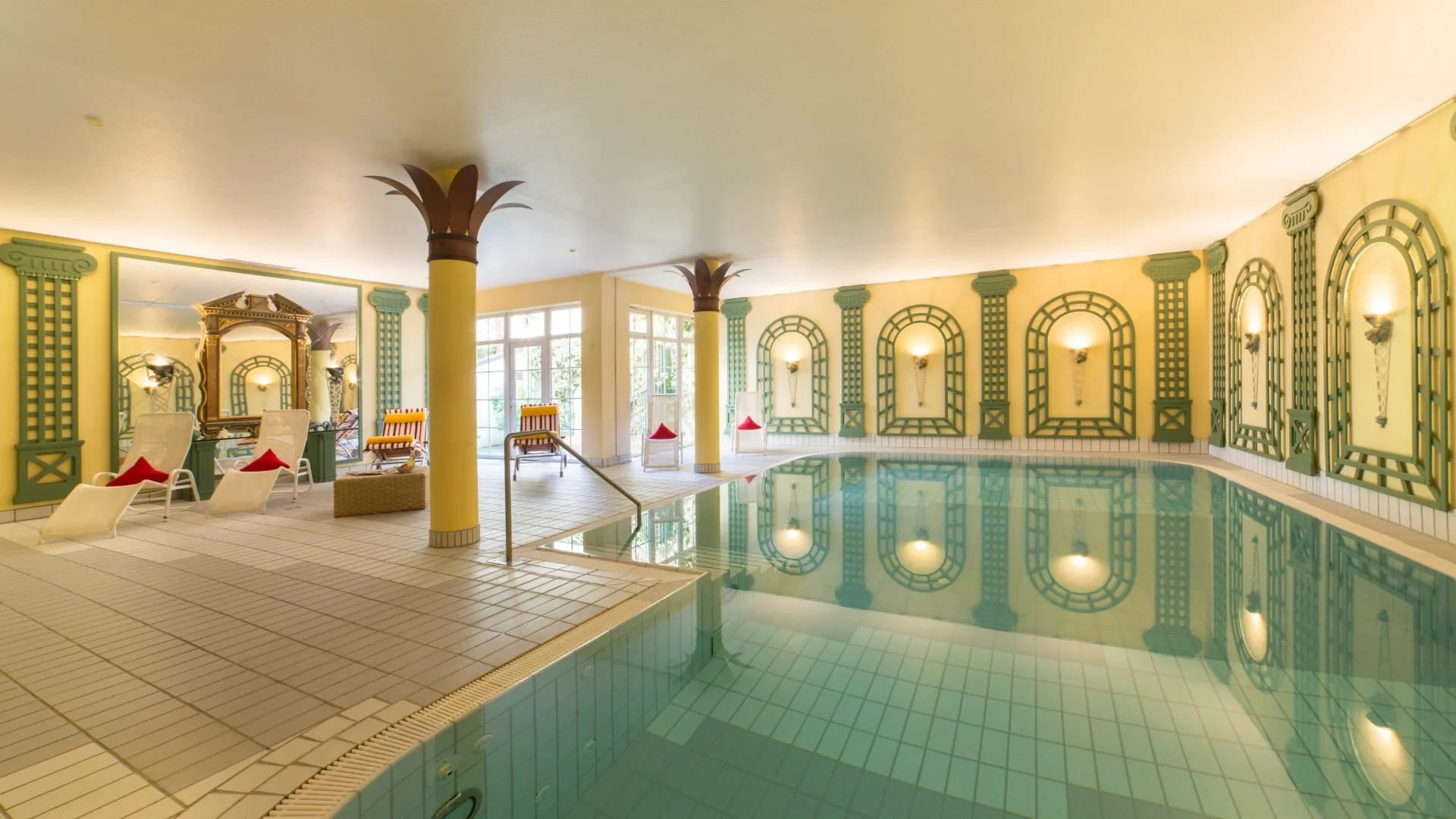 Hallenbad mit türkisem Becken, Säulen mit Palmenkapitellen und Liegen am Beckenrand