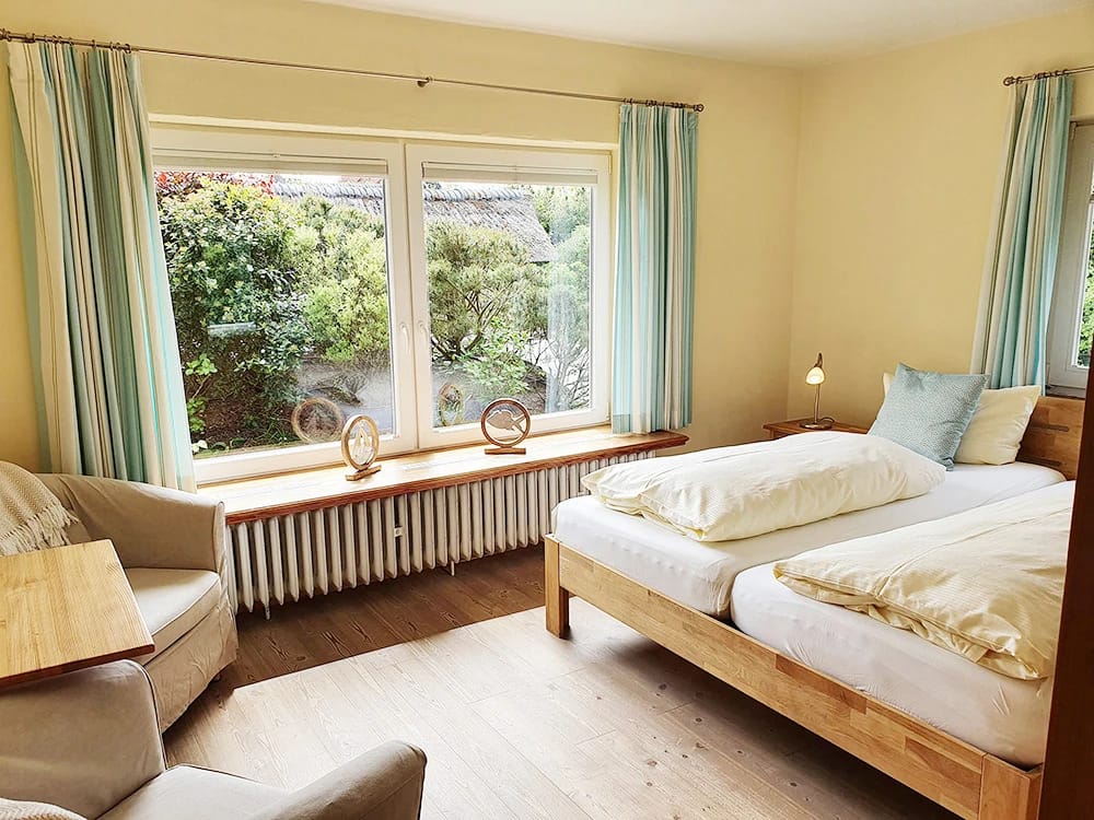 Zimmer mit zwei Betten, türkisfarbenen Vorhängen, Sessel und Fenster zum Garten