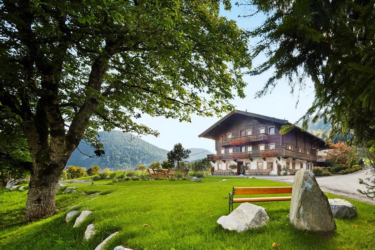 Tiroler Landhaus mit Holzbalkonen, davor Wiese mit großem Laubbaum und Sitzbank