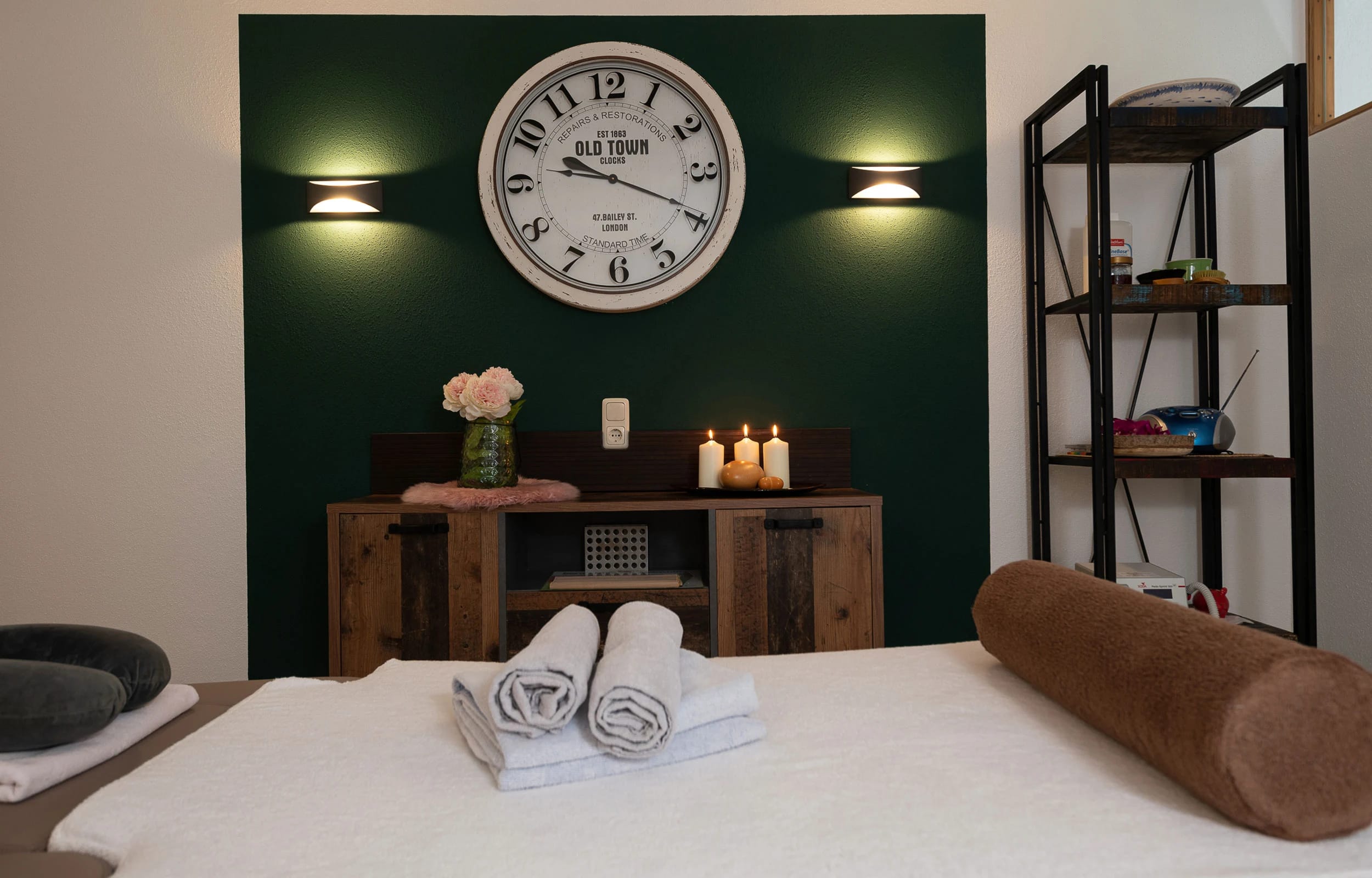 Massageraum mit Behandlungsliege, gerollten Handtüchern, dunkelgrüner Wand, Wanduhr und Kerzen