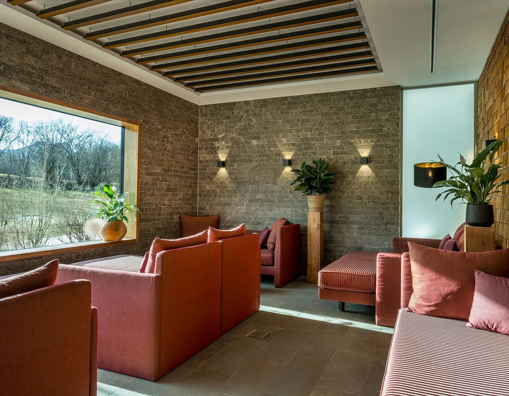 Lounge-Sitzbereich mit roten Samtsofas, Natursteinwand und Fensterfronten zum Garten