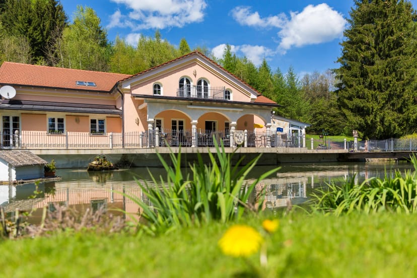 Außenansicht des lachsfarbenen Haupthauses mit Säulenterrasse, Gartenteich und Wald im Hintergrund