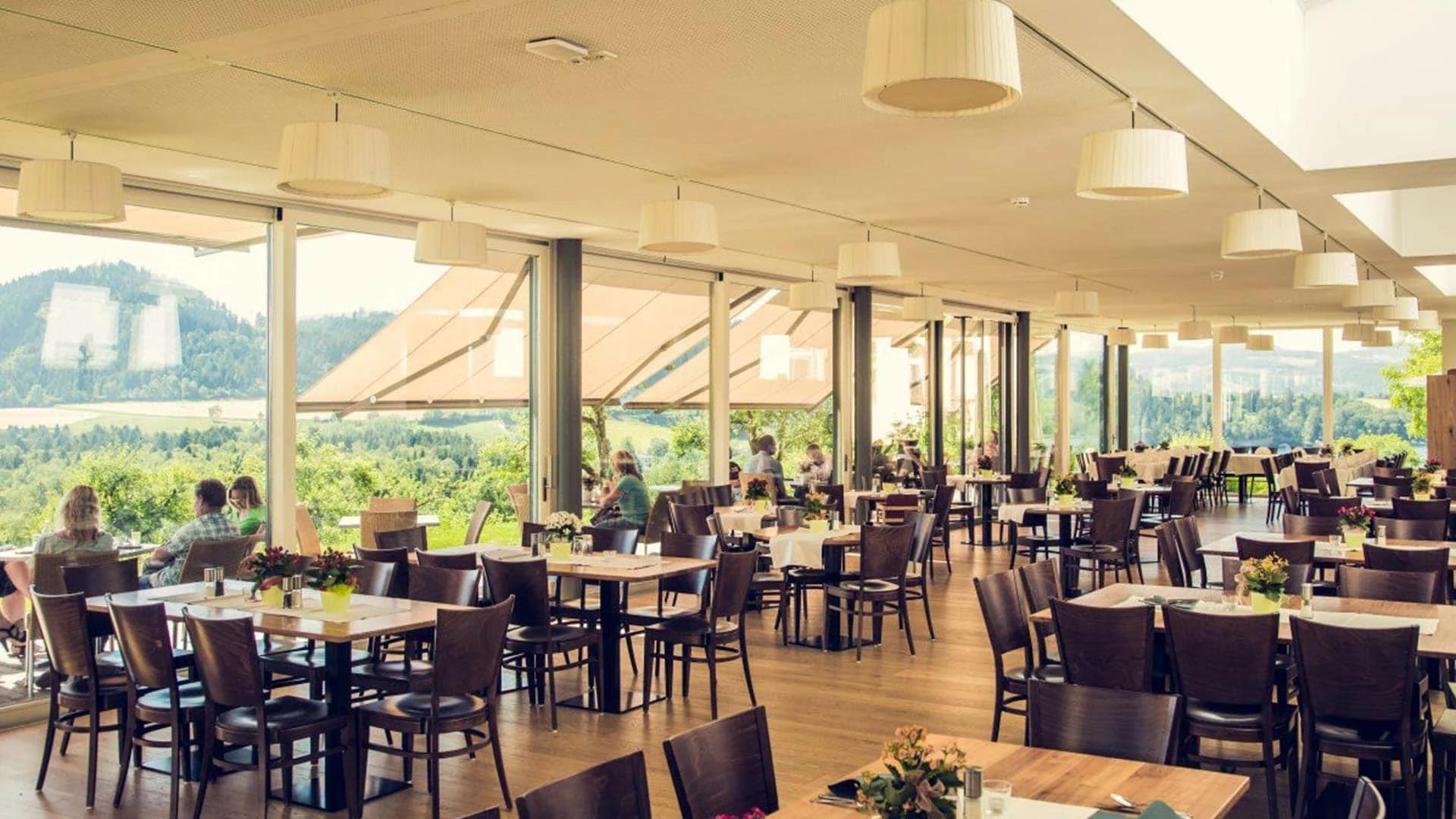 Heller Restaurantsaal mit Pendelleuchten, Holzstühlen und Panoramafenstern mit Blick ins Grüne